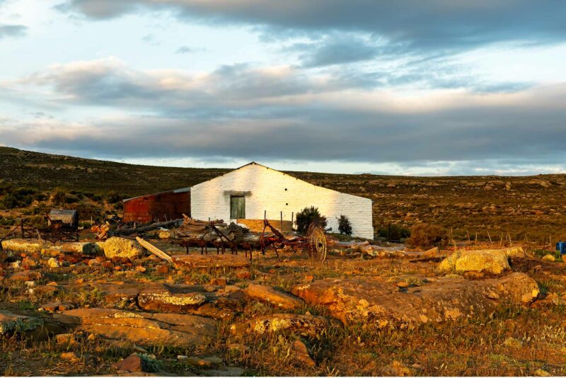 Winter Skies In Sutherland – Original Nikon photography and copyright by CFP Prints & Craig Fouché Photography. A rusty tractor rake leads to a white Karoo farmhouse glowing in soft sunset light beneath a moody winter sky in Sutherland, Northern Cape, South Africa.