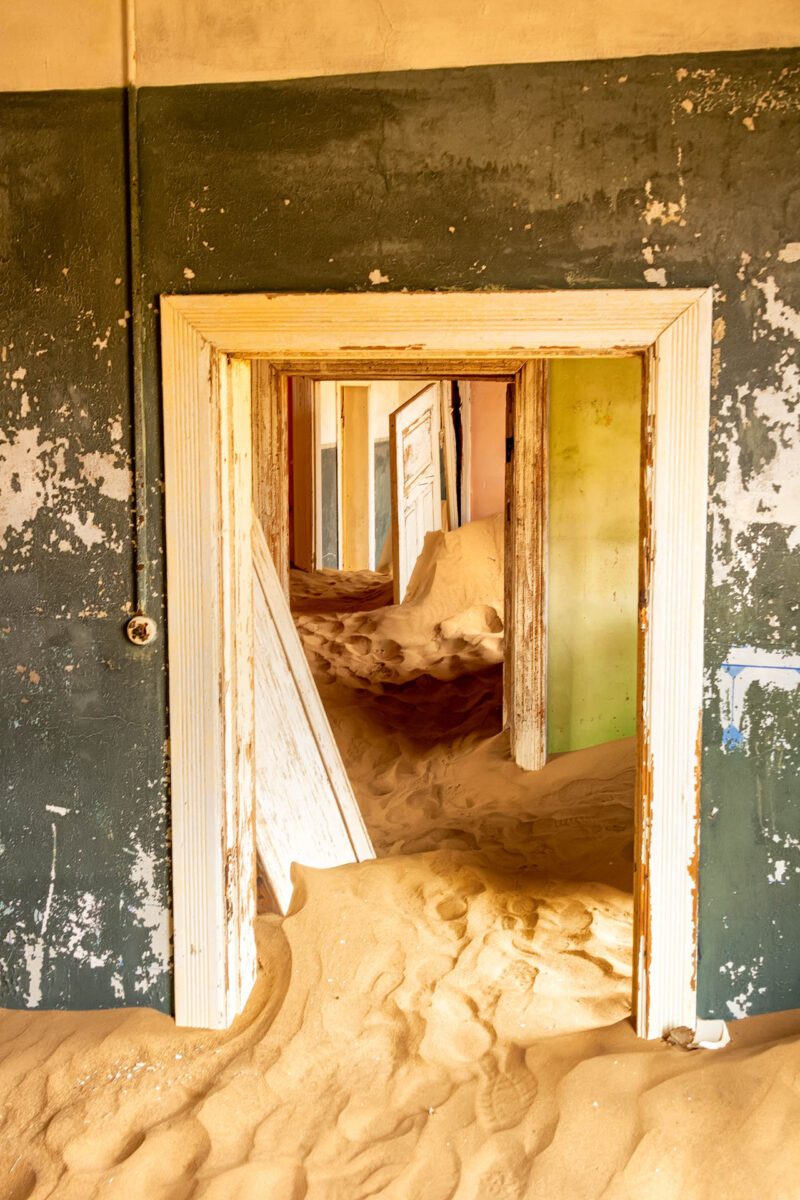 Layered doorways in a sand-filled corridor at Kolmanskop, Namibia, captured in soft golden light—fine art colour photograph of African ghost town interior.