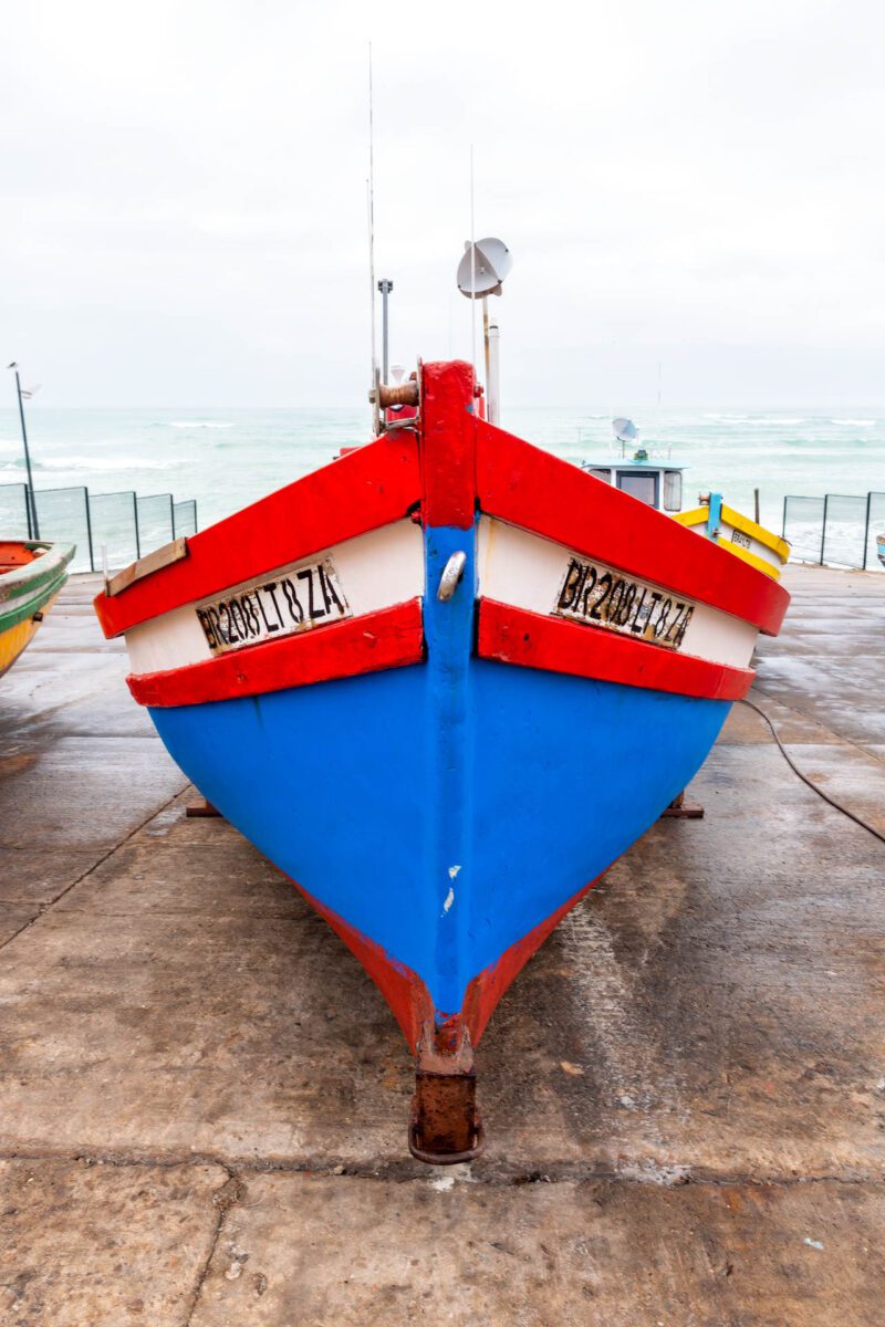 Red, blue, and white trawler beached on Arniston slipway under grey stormy skies, symbolising calm and spiritual anchoring in South Africa. Original Nikon photography and copyright by CFP Prints & Craig Fouché Photography