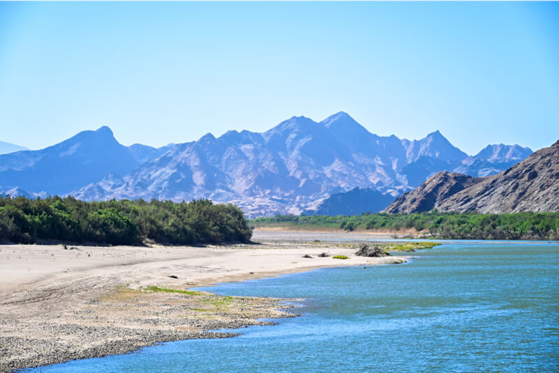 Orange River flowing past a sandy bank and green vegetation beneath the rugged Richtersveld mountains on the Namibian border